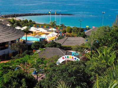 Blick auf eine bunte Ferienanlage mit Pool, Sonnenschirmen und blauem Meer im Hintergrund.