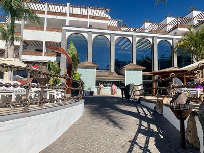 Hotel s bazénem, slunečníky a modrou oblohou pod zastřešenou terasou s velkými okny.