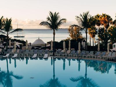 Großer Pool mit Liegestühlen und Palmen, im Hintergrund Blick auf Meer und Berge bei Sonnenuntergang.
