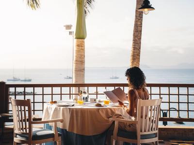 Žena sedí u prostřeného stolu s výhledem na moře při západu slunce.