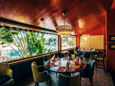 Restaurant élégant avec plafond en bois, grandes fenêtres et vue sur la piscine et les palmiers.