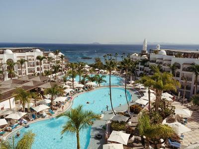 Grand complexe avec plusieurs piscines, palmiers et vue sur la mer sous un ciel clair.