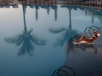 Person entspannt sich auf einer Leiter im großen, ruhigen Hotelpool mit Palmenreflexion.