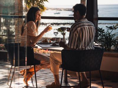 Zwei Personen sitzen bei Sonnenuntergang in einem Café mit Meerblick und unterhalten sich.