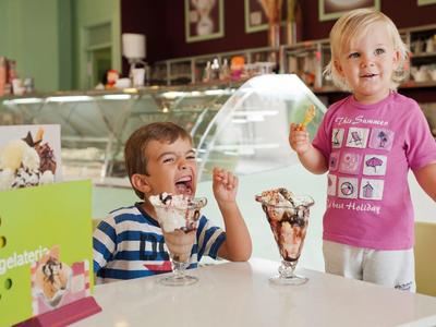 Zwei lachende Kinder mit Eisbechern sitzen an einem weißen Tisch in einer Eisdiele.