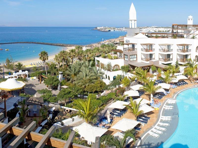 Complexe hôtelier avec grande piscine, transats, palmiers et mer en arrière-plan sous un ciel clair.