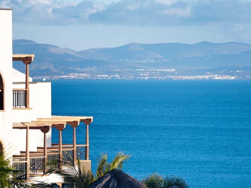 Vue sur la mer depuis une terrasse avec un bâtiment méditerranéen et des montagnes en arrière-plan.