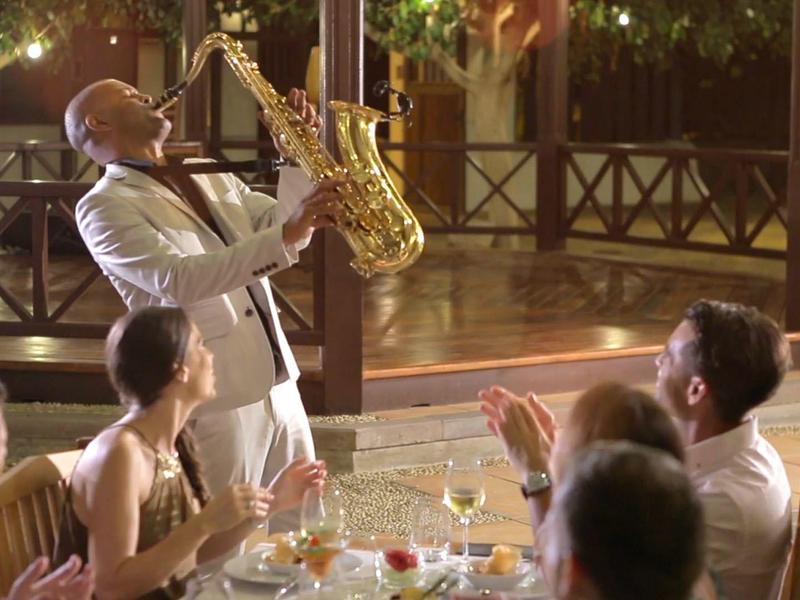 Un saxophoniste joue pour des invités lors d'un dîner en plein air, entouré d'éclairages décoratifs.