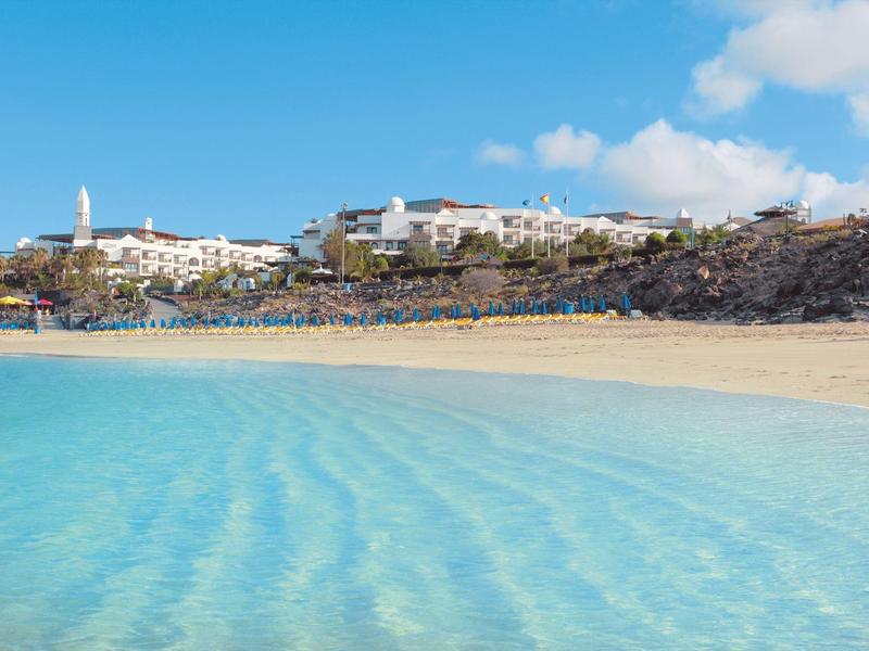 Eau turquoise claire avec plage de sable et bâtiments sur une colline rocheuse sous un ciel bleu