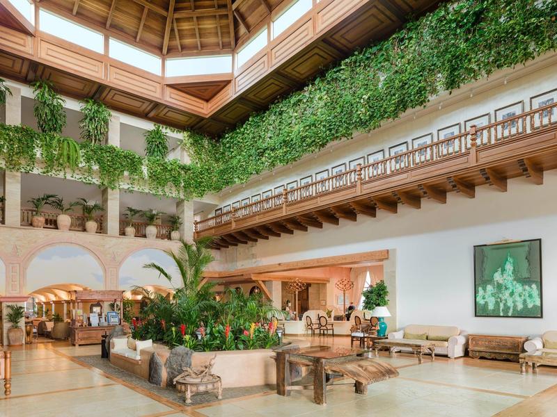 Hall d'hôtel lumineux avec des plantes vertes luxuriantes et des espaces de détente sous un plafond à poutres en bois.