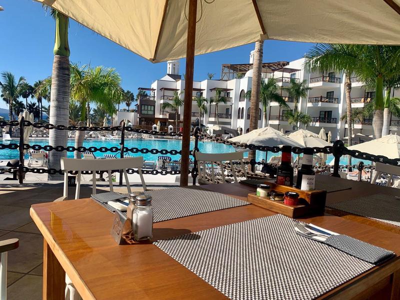 Coin repas extérieur avec table en bois, parasol et vue sur la piscine et les bâtiments de l'hôtel.