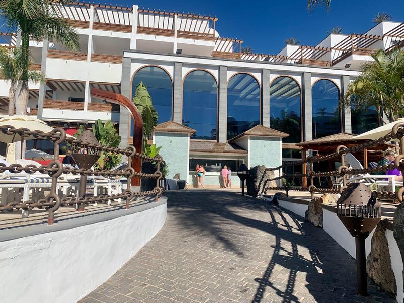Entrée d'un hôtel avec de grandes arches en verre et des palmiers sous un ciel ensoleillé.