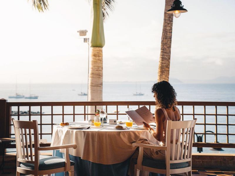 Femme assise à une table avec une nappe sur une terrasse donnant sur la mer au coucher du soleil.