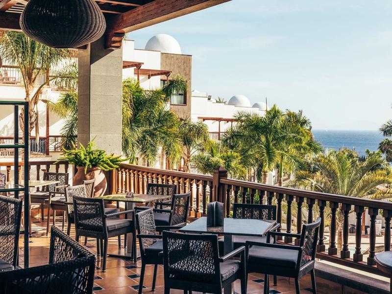 Terrasse d'hôtel avec tables et chaises, entourée de palmiers et vue sur l'océan.