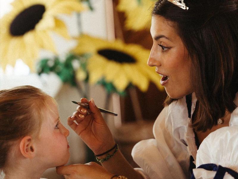 Une femme portant une tiare maquille une jeune fille à côté d'une table avec des tournesols.