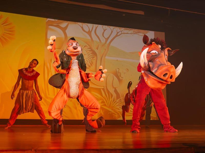 Spectacle sur scène avec les personnages Disney Dingo et Pumba en costumes de safari devant un fond jaune.