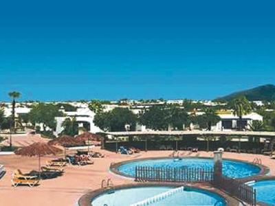 Schwimmbad mit blauen Pools, Liegestühlen und weißen Gebäuden vor bewölktem Himmel.