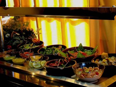 Buffet mit verschiedenen Salaten, Garnelen und Saucen vor hellgelten Vorhängen.