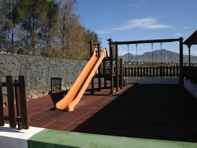 Spielplatz mit orangener Rutsche, Holzzäunen und grauer Steinmauer im Hintergrund bei Sonnenschein.
