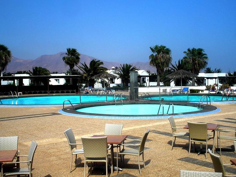 Round pool with sun loungers and tables surrounded by palm trees and hotel buildings.