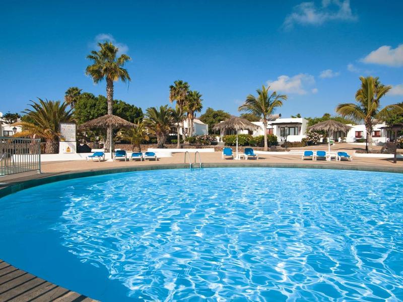 Large pool with blue water surrounded by sun loungers and palm trees under clear sky.