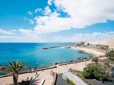 Küste mit blauem Meer, Palmen, Promenade und Gebäuden unter bewölktem Himmel.