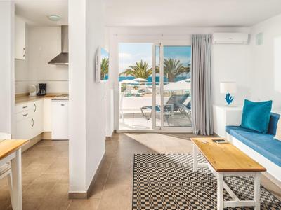 Helles Wohnzimmer mit weißer Einrichtung, blauen Kissen, Holz- und Rattanmöbeln, Blick auf Terrasse und Meer.