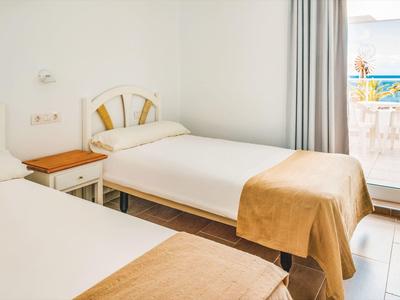 Zweibettzimmer mit weißem Möbel, hellen Wänden, braunen Decken und Blick auf Balkon mit Meer.