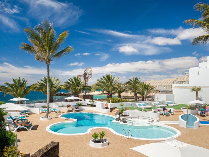 Hotel pool area with umbrellas, loungers, palm trees, and sea view under a blue sky.