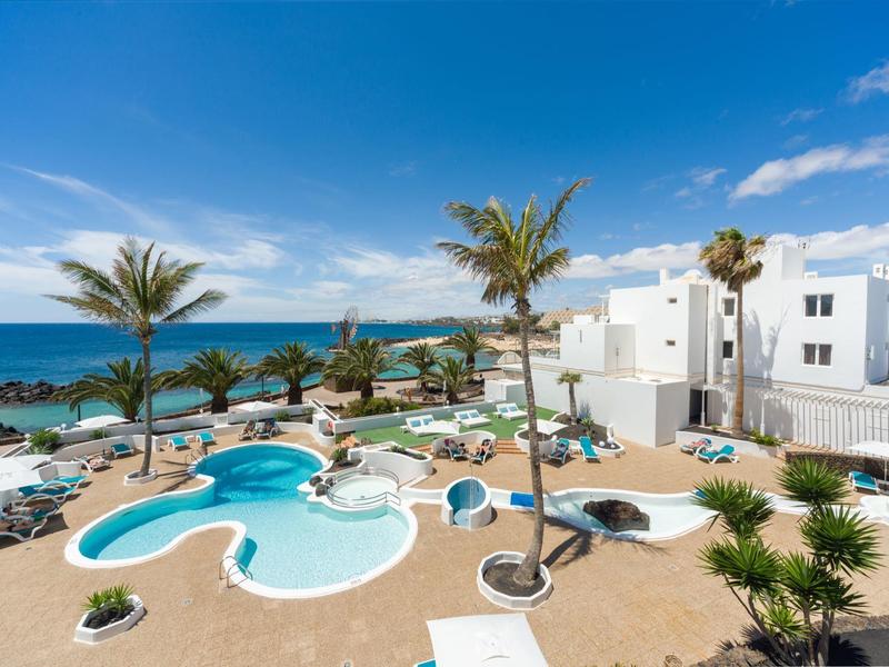 Hotel pool with palm trees and sea view under a blue sky.