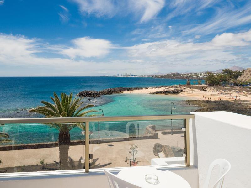 View from a balcony overlooking a beach with clear water and palm trees under a blue sky.