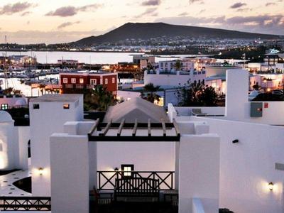 Bílé budovy s terasami s výhledem na město a horu při západu slunce.