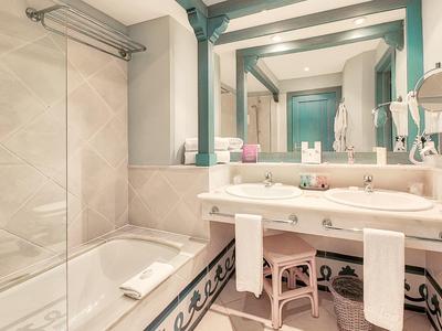 Světlná koupelna s vanou, velkým zrcadlem a dvojitou umyvadlem s ručníky a stoličkami.