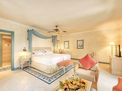 Světlý hotelový pokoj s velkou postelí, křesly, stropním ventilátorem a květinovou výzdobou.