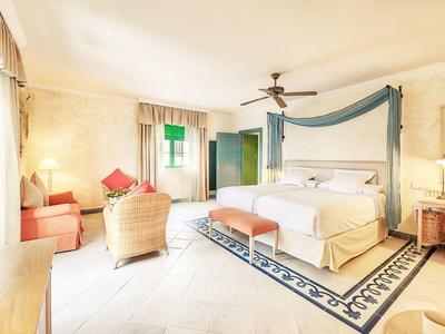 Světý hotelový pokoj s postelí s baldachýnem, křesly a stropním ventilátorem.