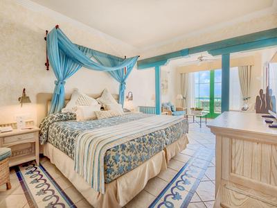 Světý hotelový pokoj s postelí s baldachýnem, vzorovaným kobercem a balkónem s výhledem na moře.