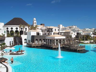 Rezort s velkým bazénem, fontánou a bílými budovami pod jasně modrou oblohou.