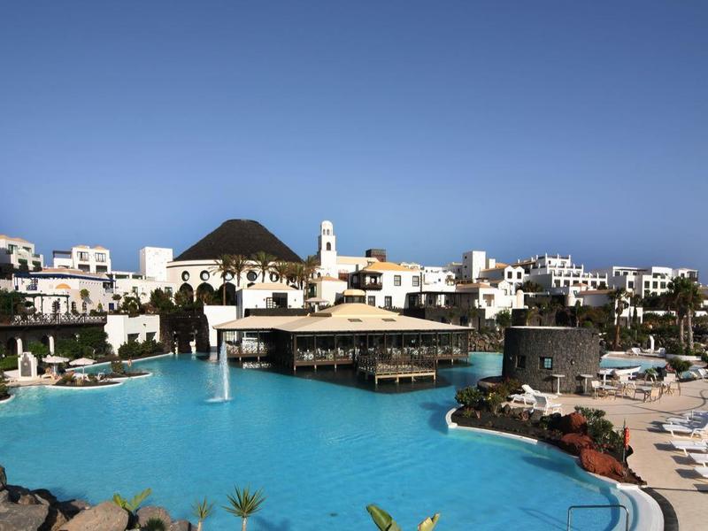 Velký venkovní bazén s lehátky kolem, obklopený bílými budovami pod jasnou modrou oblohou.