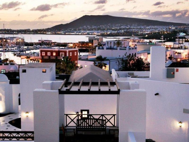 Bílé budovy s terasami s výhledem na město a horu při západu slunce.
