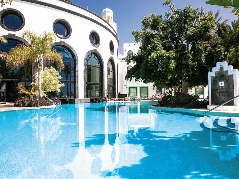 Zakřivená fasáda hotelu s velkými okny vedle průzračného modrého bazénu obklopeného stromy.