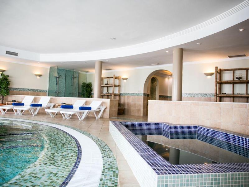 Vnitřní bazén s lehátky a malým vířivým bazénem v moderním lázeňském zařízení.