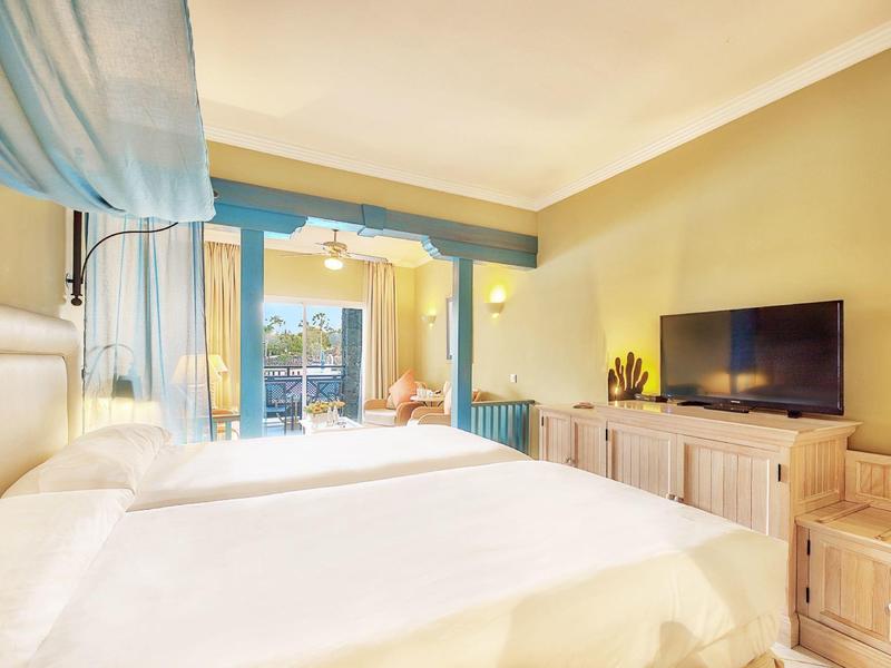 Světý hotelový pokoj se dvěma postelemi, TV a balkonem s venkovním posezením.