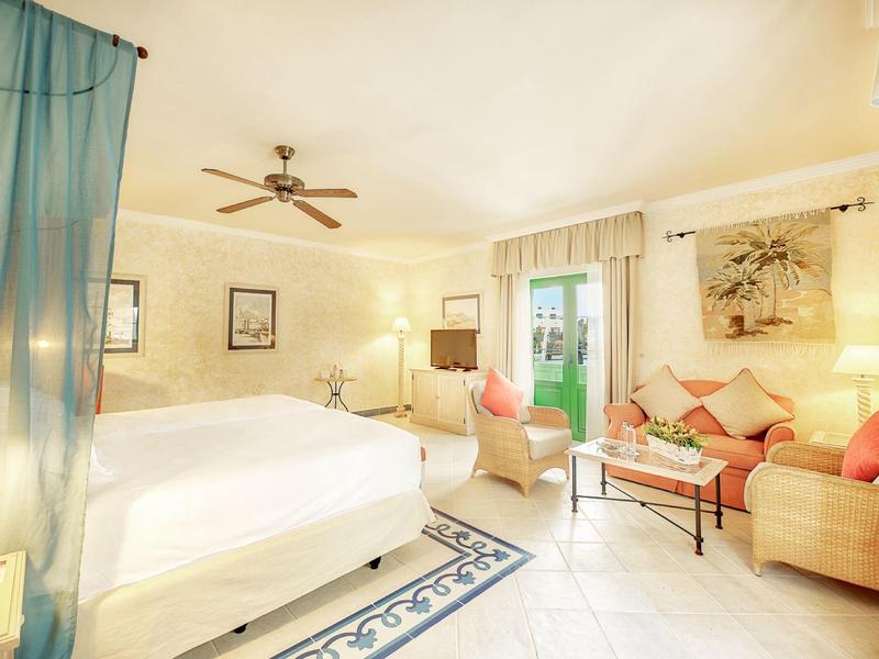 Světý hotelový pokoj s postelí, pohovkou, konferenčním stolkem a balkónovými dveřmi v teplých tónech.