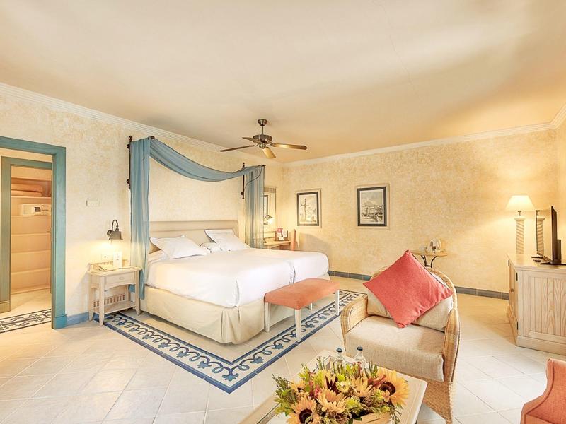 Světlý hotelový pokoj s velkou postelí, křesly, stropním ventilátorem a květinovou výzdobou.