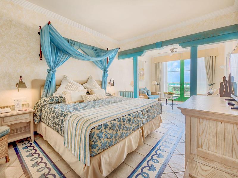 Světý hotelový pokoj s postelí s baldachýnem, vzorovaným kobercem a balkónem s výhledem na moře.