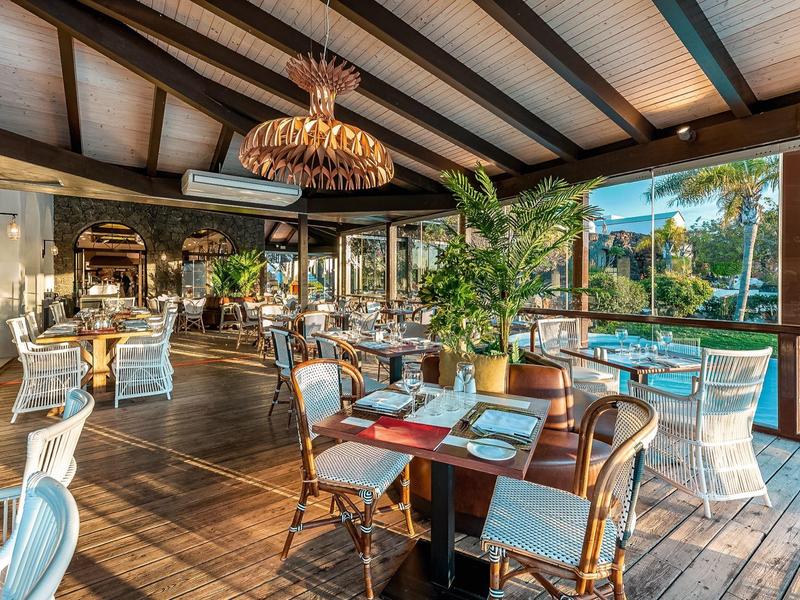 Příjemná hotelová jídelna s dřevěnými trámy, tropickými rostlinami a velkými okny s výhledem na bazén.