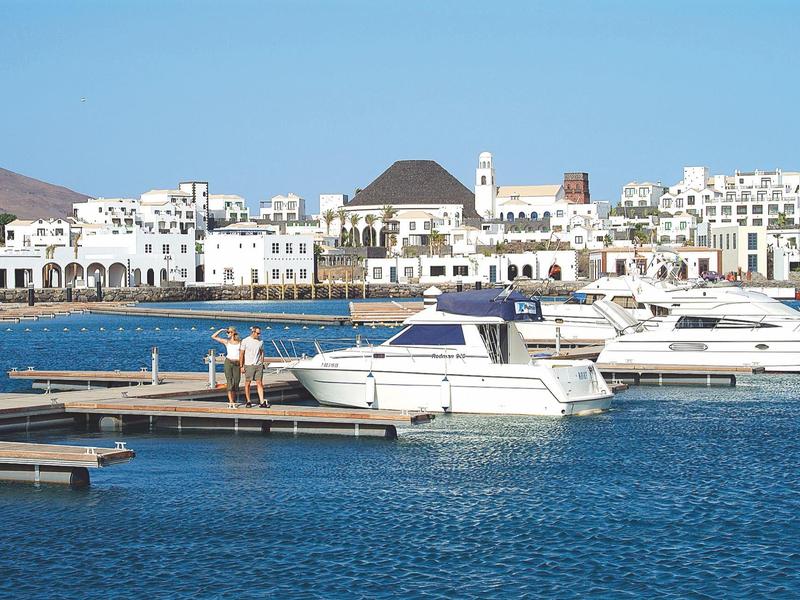 Pohled na bílé budovy u maríny s jachtami a klidnou modrou vodou pod jasnou oblohou.