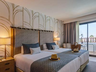 Camera d'hotel moderna con due letti, comodini, lampade e balcone con vista sulle palme.