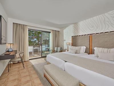 Camera d'hotel luminosa con due letti, scrivania, TV e balcone con vista sugli alberi.