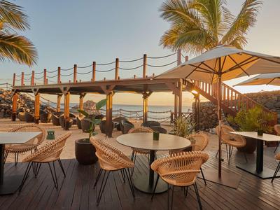 Área exterior de un hotel con mesas, sillas y una terraza de madera junto al mar al atardecer.